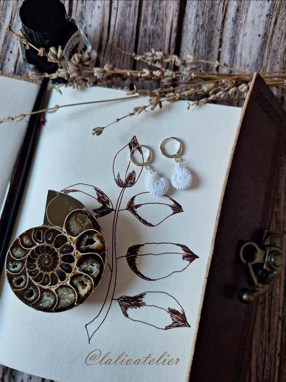 Mikrogehäkelte Ammoniten-Creolen im Vintage-Stil dekoriert mit Notizbuch, Fossil, Lavendel und Füllfederhalter auf dunklem Holz