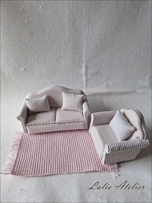 Two miniature sofas with beige stripes stand on a hand-woven, rose-coloured miniature rug. The scene is set against a neutral fabric background and features intricately detailed doll’s house accessories from Lalio Atelier