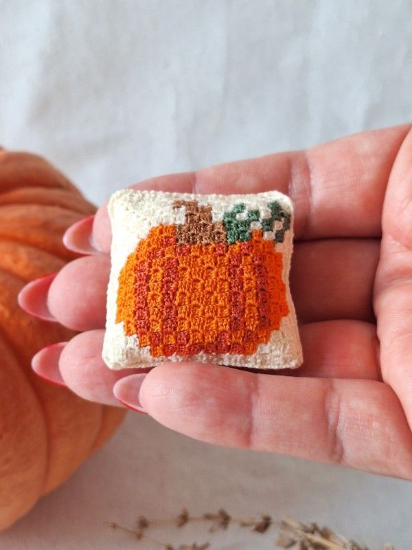 Handmade micro crochet pumpkin cushion held between fingers showing its miniature size